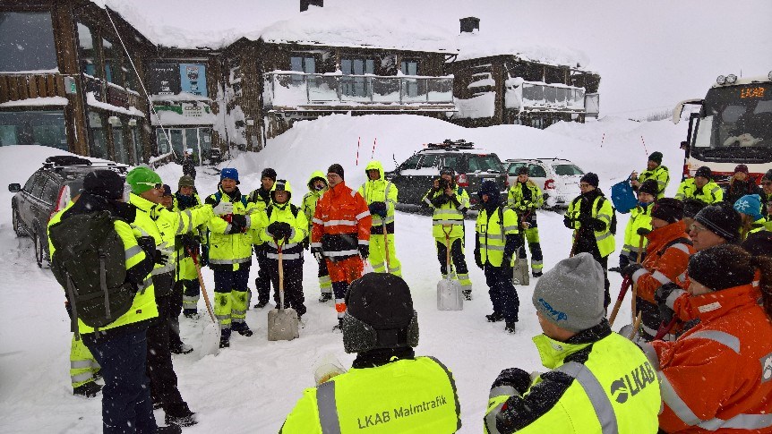 En grupp människor i LKAB-kläder och med snöskyfflar i händern samlas i Katterjåkk. a