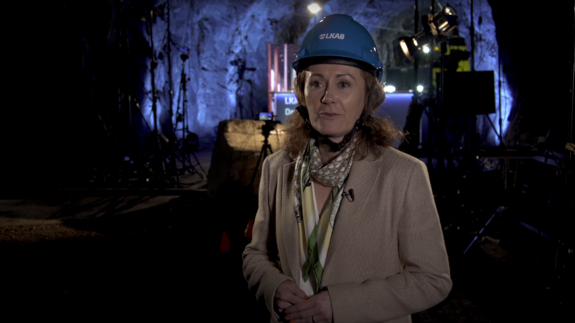 Kvinna i gruvhjälm, under jord, med tv-studio i bakgrunden. 