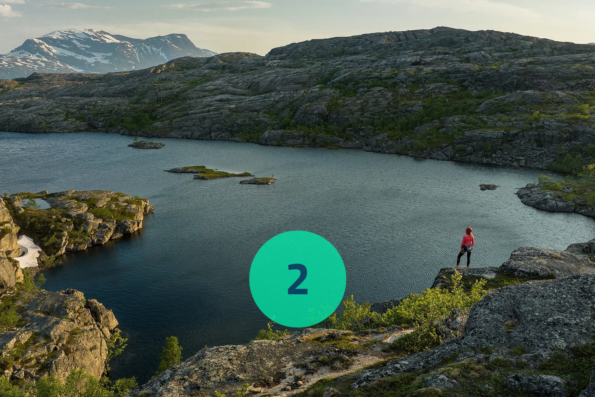 Woman in climbing gear looking out over body of water with snow capped mountains in background.