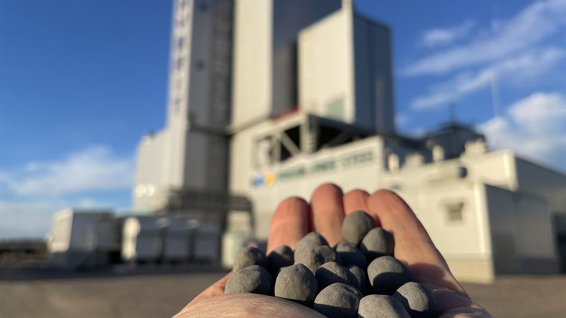 Hand med järnmalmspellets med en fabriksbyggnad i bakgrunden