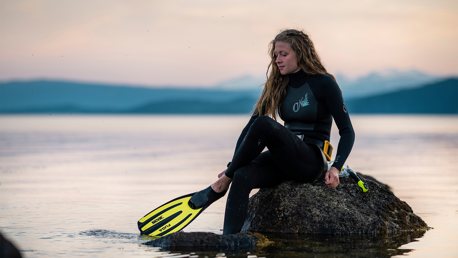 Kvinna i dykarutrustning sitter på sten i hav/sjö