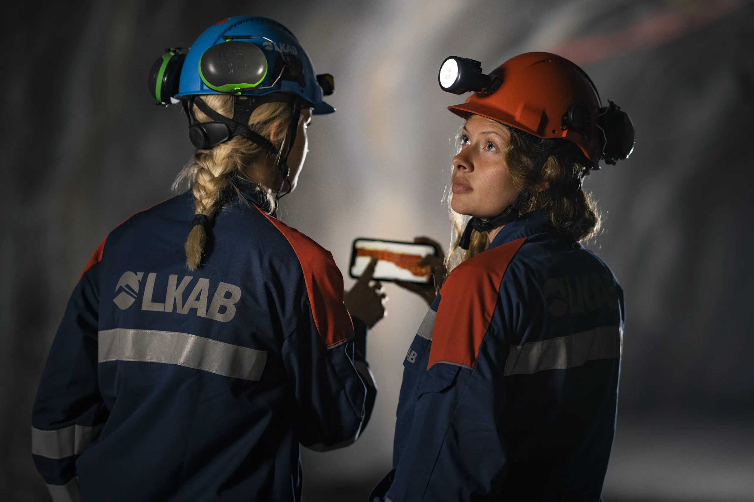 Två kvinnor i gruvkläder i underjordsmiljö håller i smart telefon.